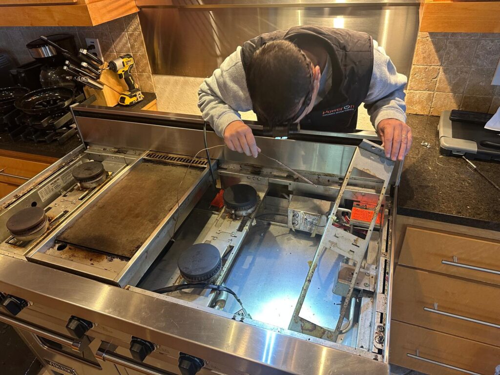 The image shows a technician working on a stove. They are inspecting the inner components of the stove, likely troubleshooting or repairing the burners and electrical connections. The technician is wearing a jacket with