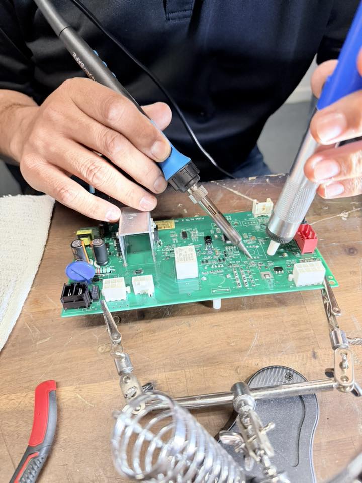 The image shows a technician soldering components onto a circuit board using a soldering iron, while holding another tool with their other hand. The circuit board is held in place with a helping hand tool for stability
