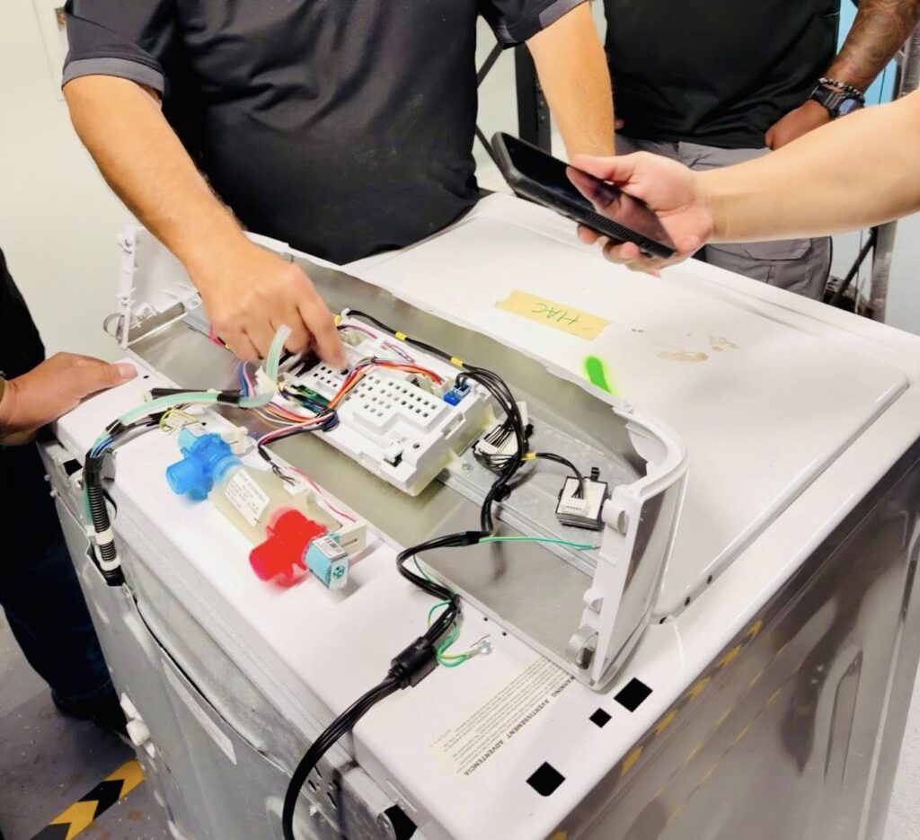 The image shows technicians working on the interior of a washing machine, with a focus on the control board and connected wiring. One technician is pointing at the wiring, while another holds a smartphone.